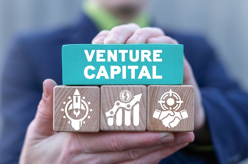 A business man holding wooden blocks, the first block printed as VENTURE CAPITAL and the other three blocks are printed with some symbols.